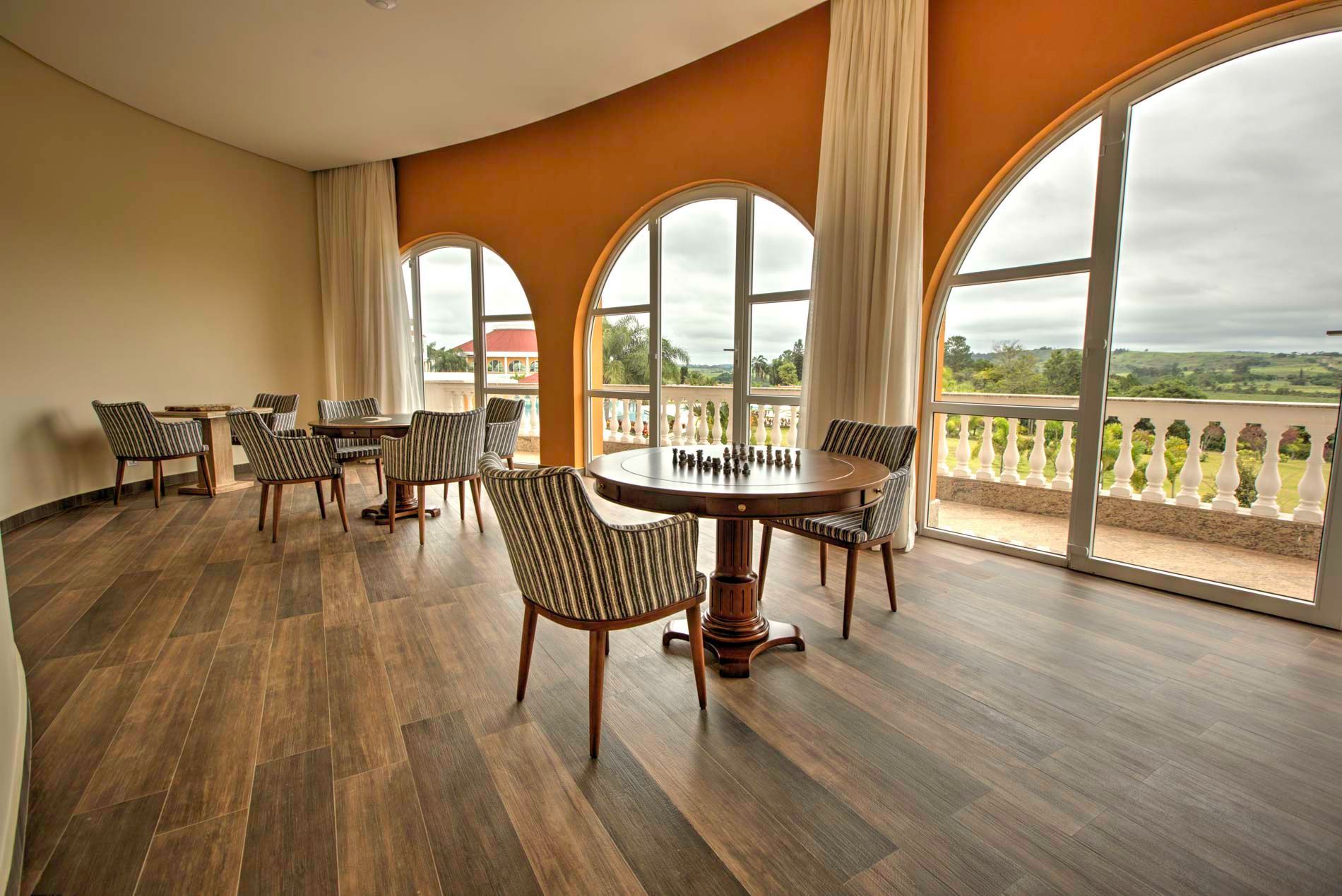 Sala com mesas, cadeiras, janelas em arco com vista para o exterior, piso de madeira clara e paredes laranja.