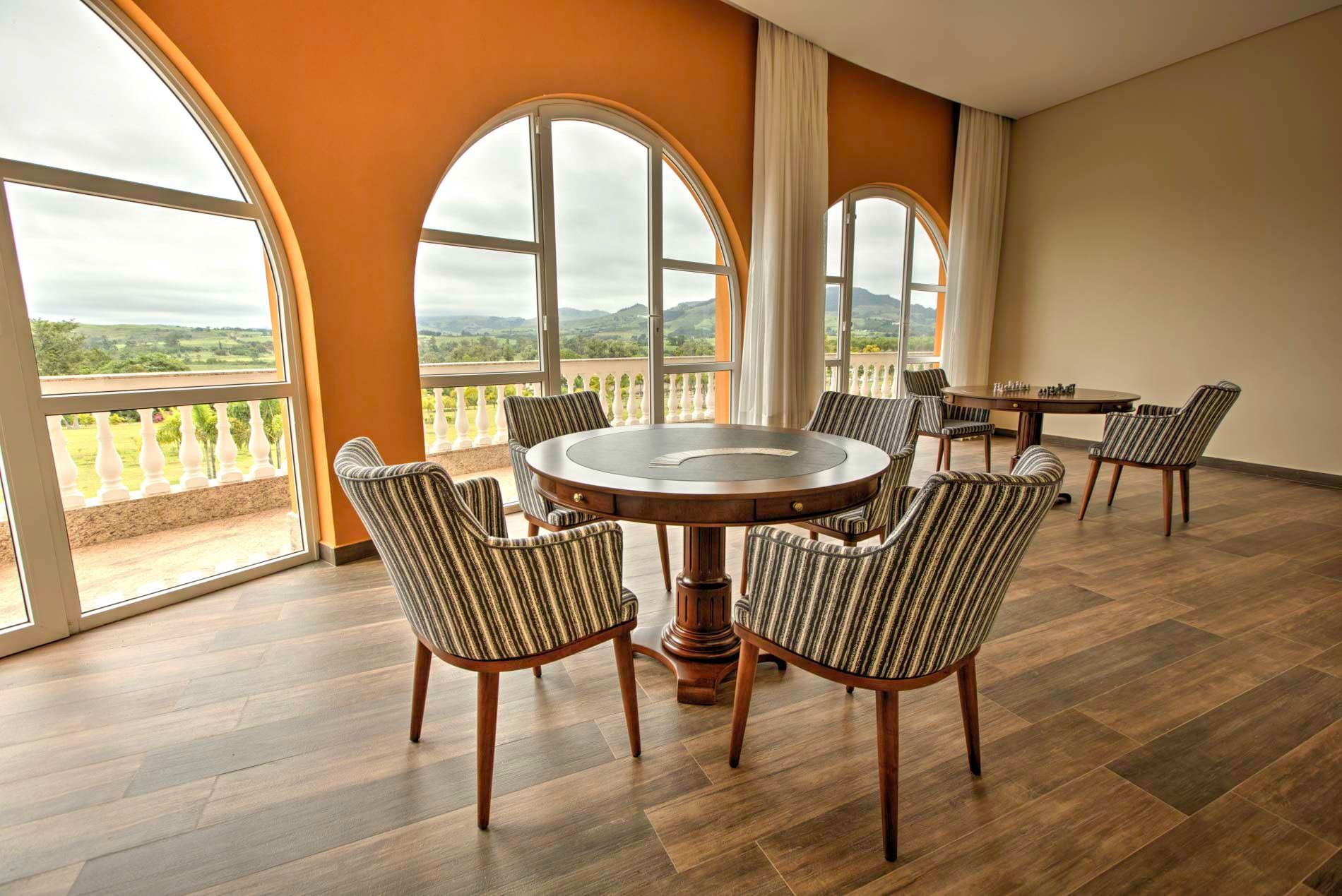Uma sala com mesa redonda e cadeiras, grandes janelas em arco com vista panorâmica, piso de madeira e paredes laranja.