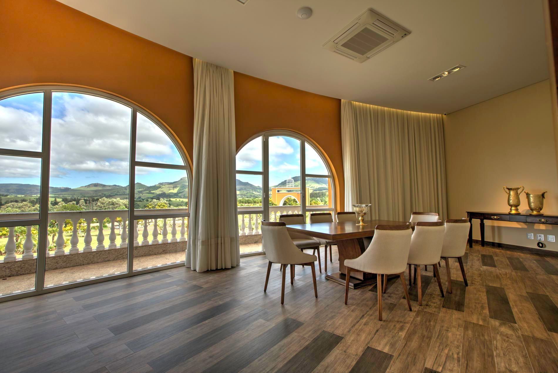 Sala de jantar com janelas em arco, mesa de madeira e cadeiras brancas, com vista para uma paisagem.