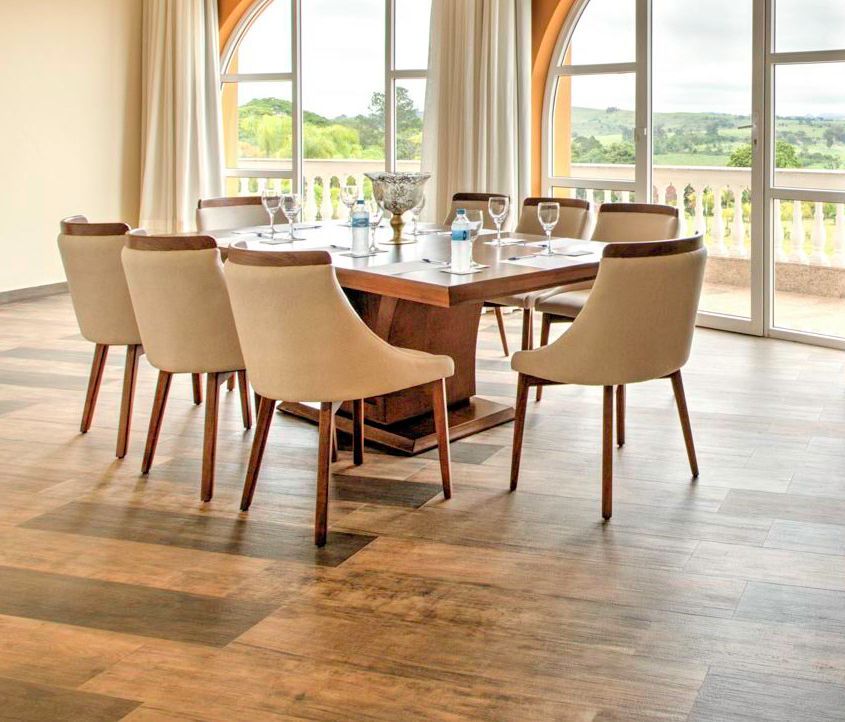 Sala de jantar com mesa quadrada de madeira, oito cadeiras e grandes janelas com vista para uma paisagem verde.