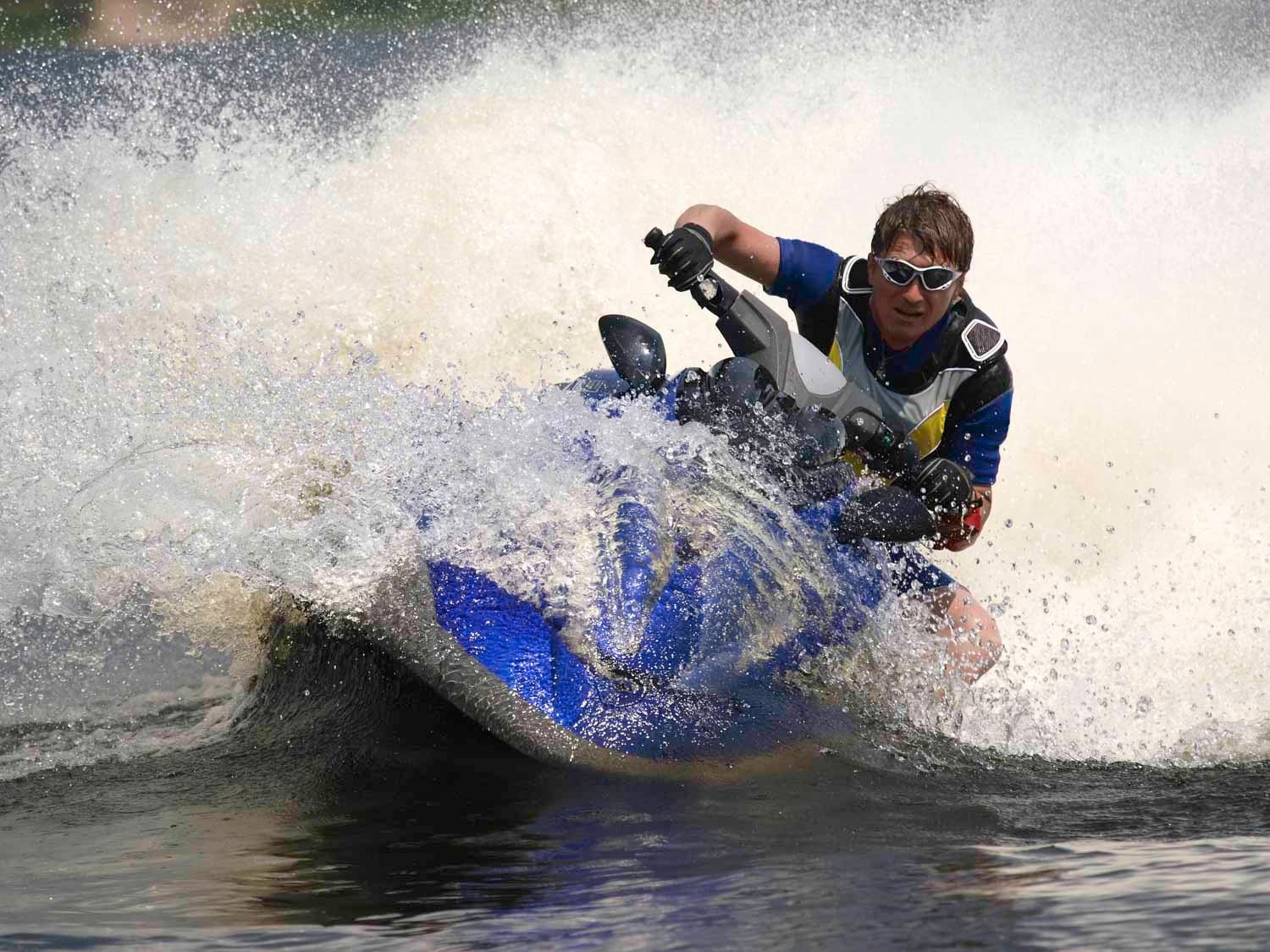 Homem em um jet ski azul, acelerando na água, causando um grande impacto.