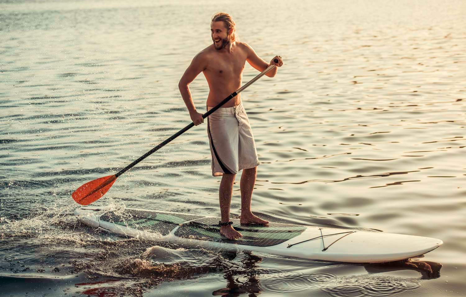 Homem pratica stand up paddle na água, segurando o remo. Ele está de shorts e sem camisa.