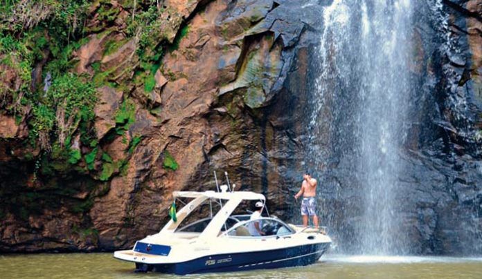 Barco perto de cachoeira com pessoa em pé no convés, sem camisa, braços levantados.