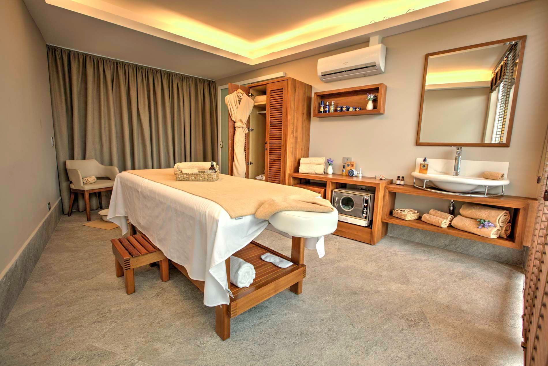 Sala de massagem com mesa de massagem, pia, detalhes em madeira e produtos de spa.