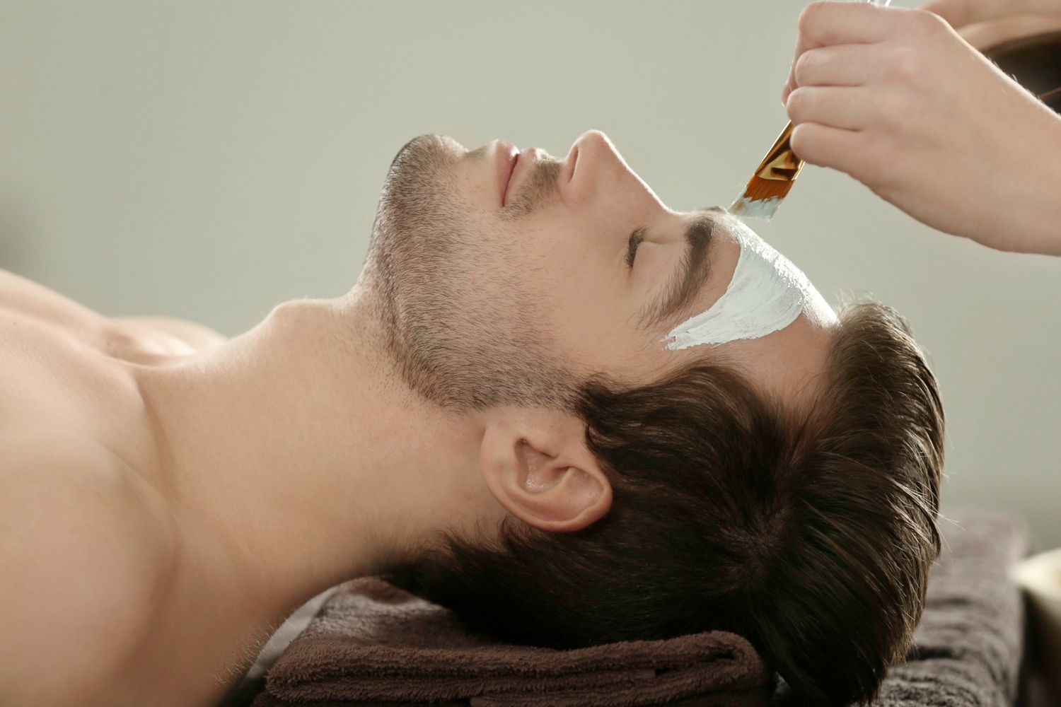 Homem recebendo uma máscara facial em um spa; creme sendo aplicado com um pincel.
