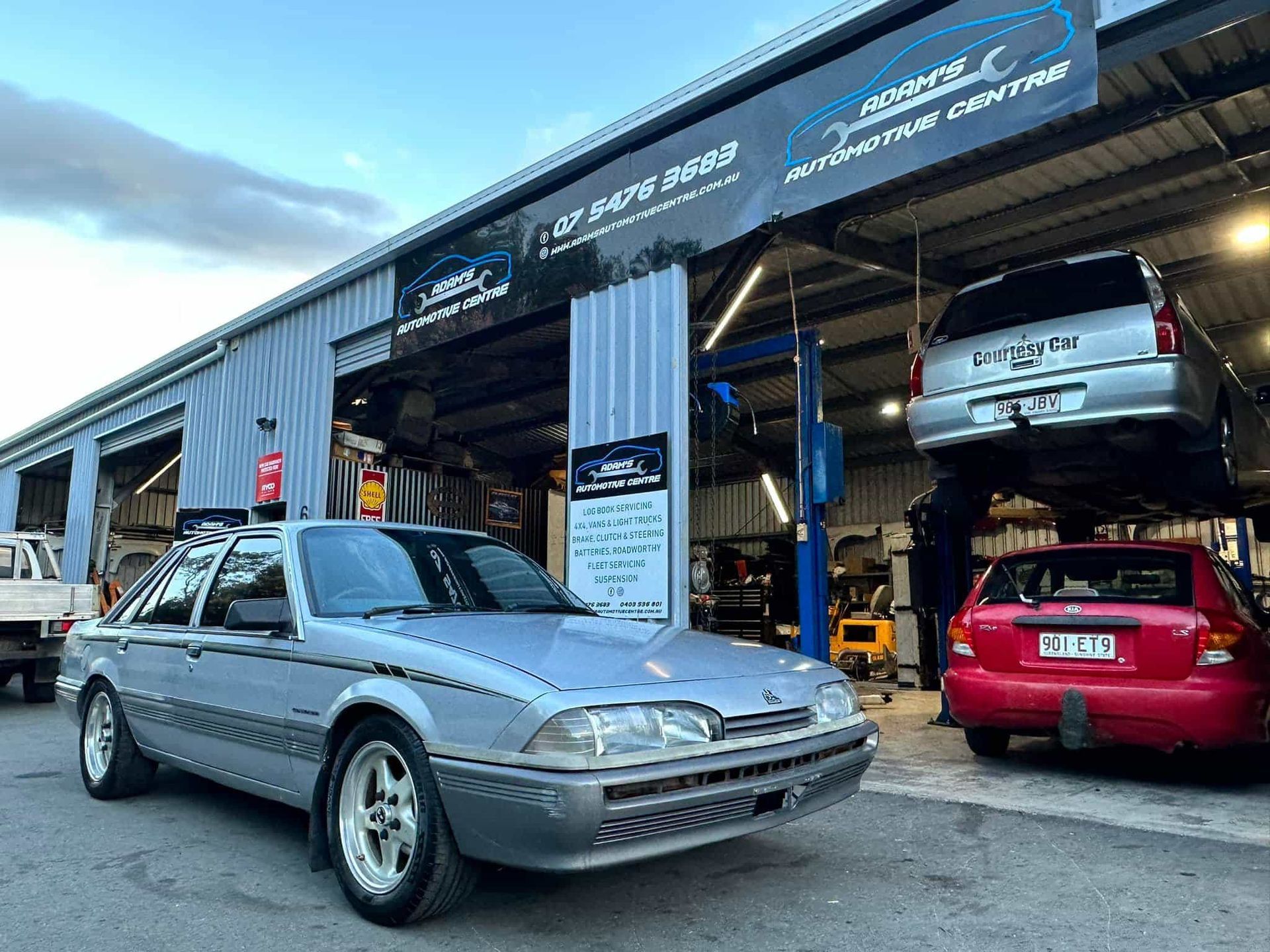 Silver car outside automotive repair shop; red car and SUV on lifts inside.  — Adam's Automotive Centre in Nambour, QLD