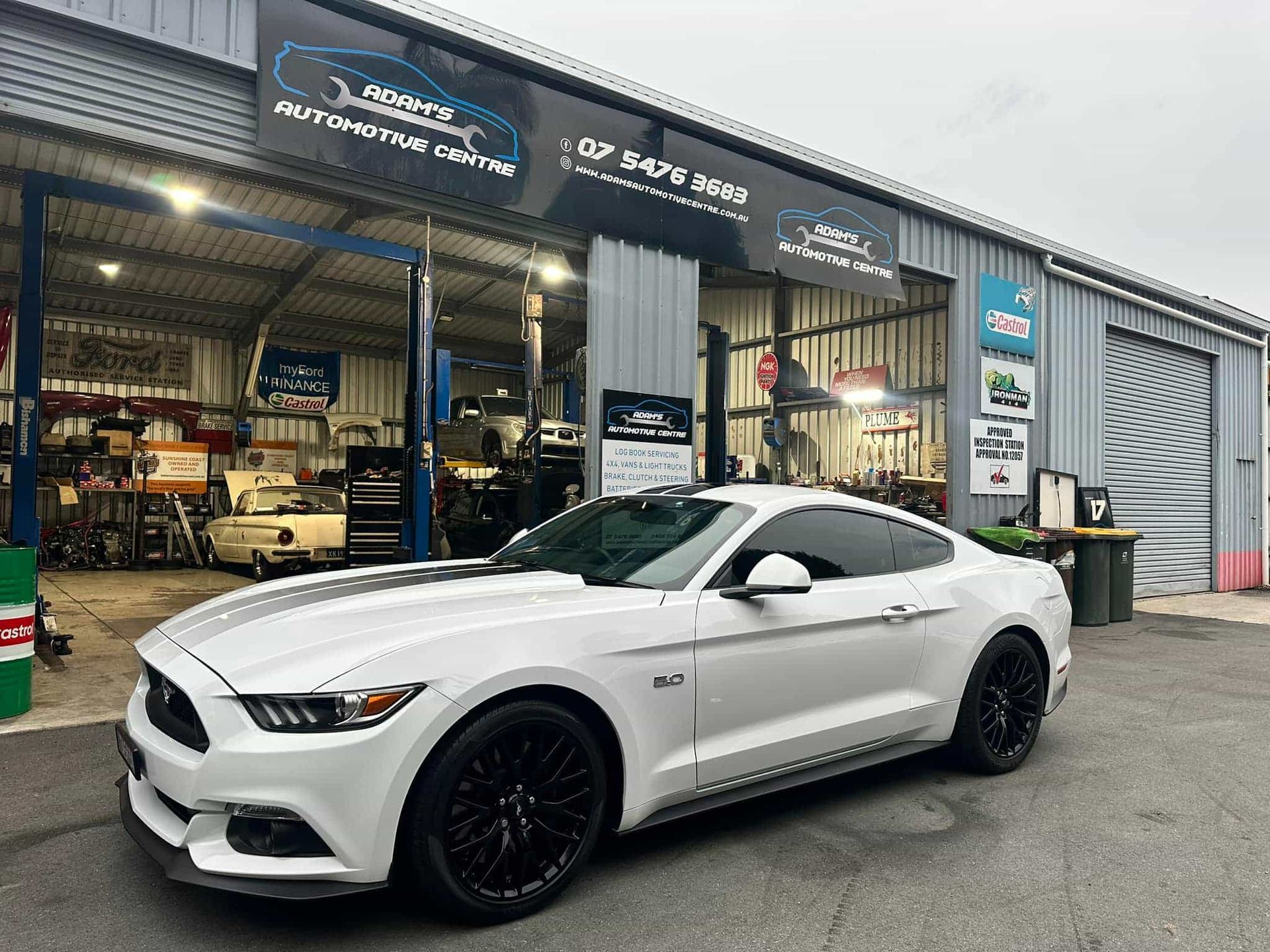 White Ford Mustang parked outside an auto repair shop with black wheels and racing stripes. — Adam's Automotive Centre in Nambour, QLD