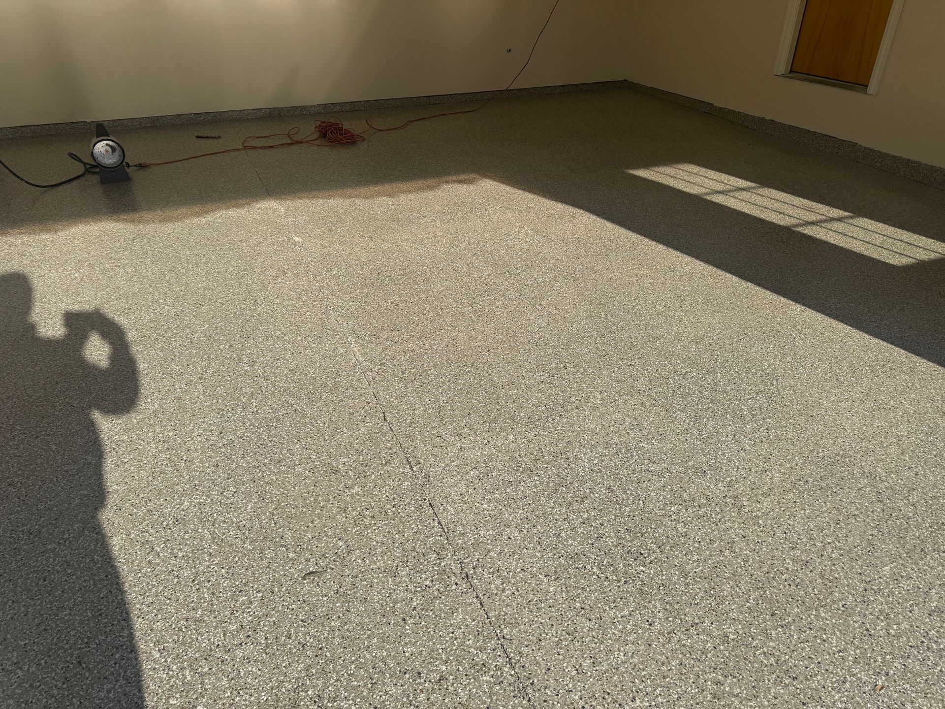Gray speckled floor, shadows of a person and sunlight across the floor. A light-colored wall is behind it.