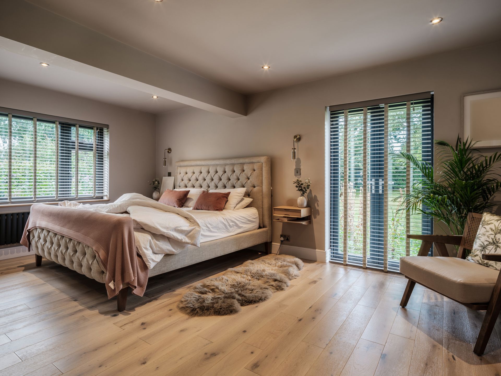 Bedroom with large bed, windows, and door to a balcony. Includes wood floors, beige walls, and a plant.