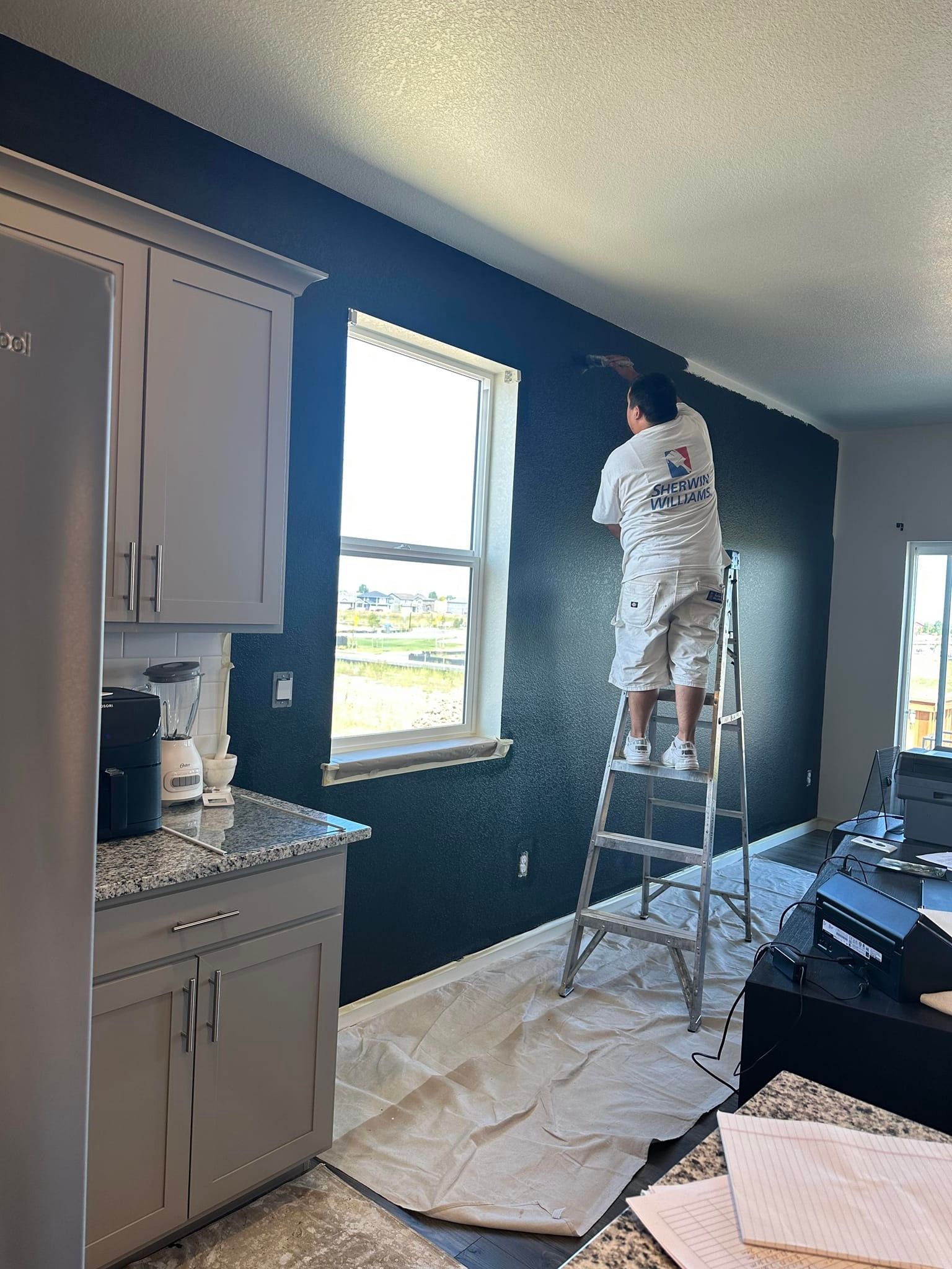 A man is standing on a ladder painting a wall in a kitchen.