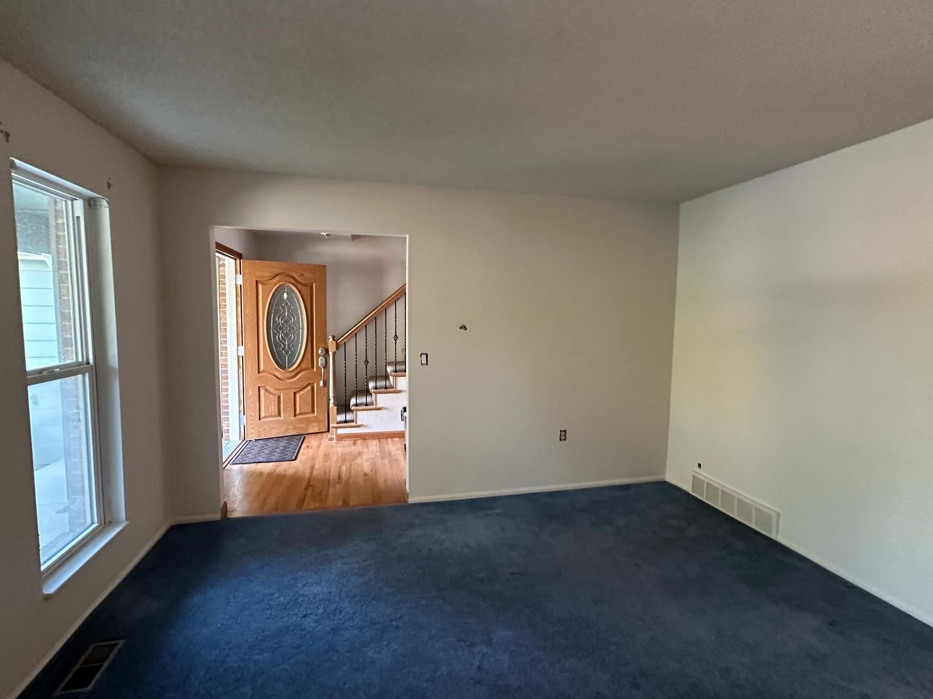 An empty living room with a blue carpet and stairs leading to the front door.