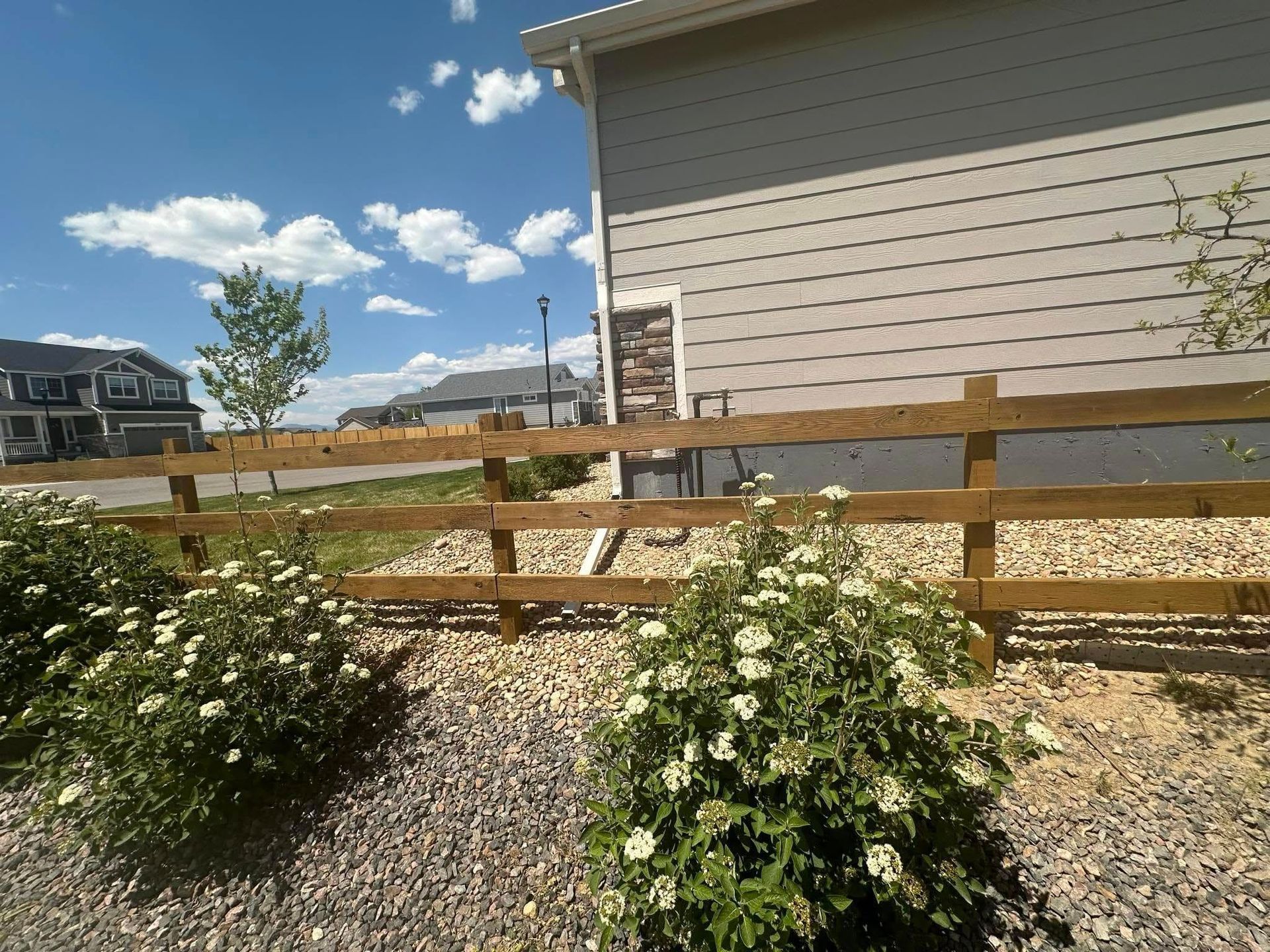 A wooden fence is surrounded by bushes and gravel in front of a house.