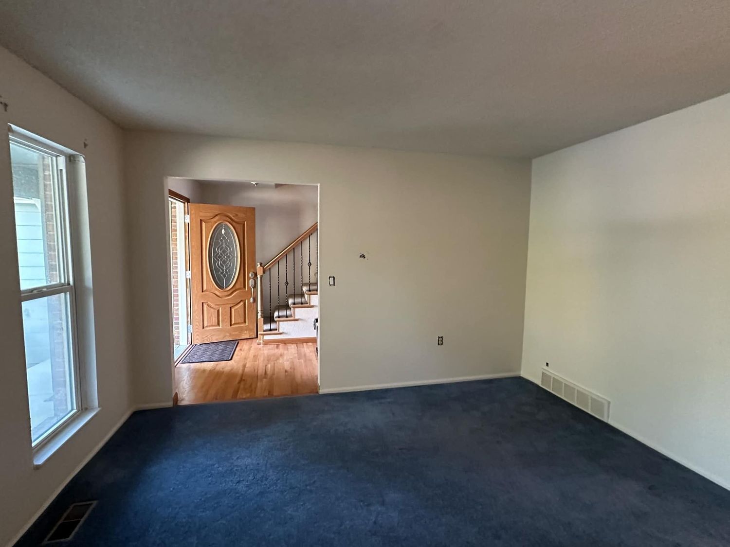 An empty living room with a blue carpet and stairs leading to the front door.