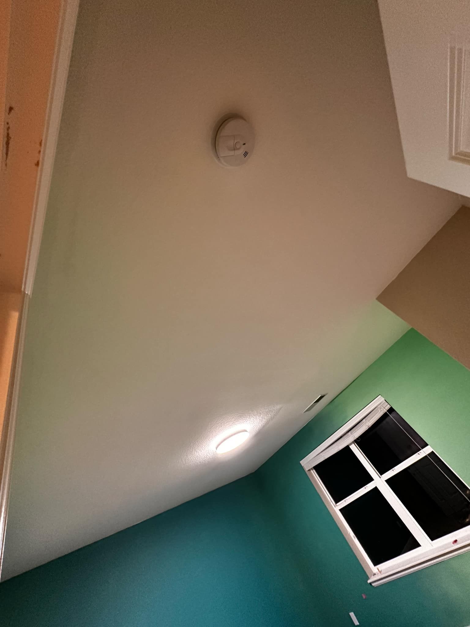 A room with a window and a smoke detector on the ceiling.