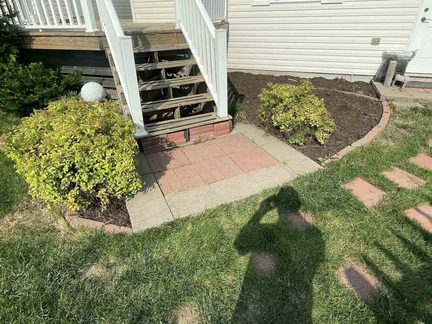 A brick and concrete pathway leads to wooden stairs next to two green shrubs and a lawn.