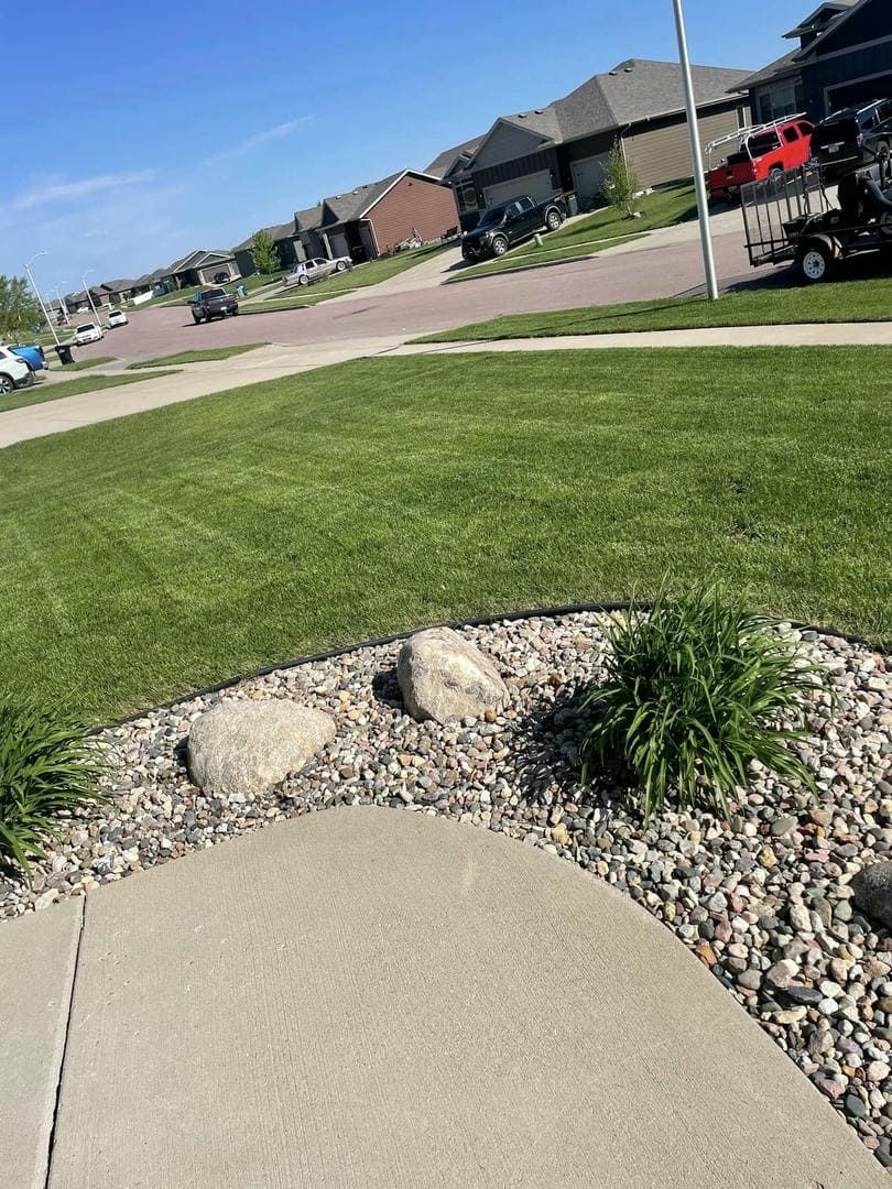 Lawn and garden bed in front of houses on a sunny day. Green grass, gray stone, and a blue sky.