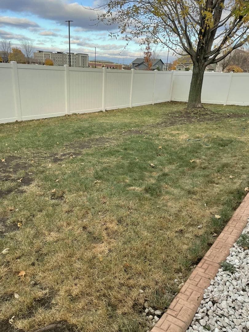 Grassy backyard with a white fence, tree, and decorative border. Overcast sky.