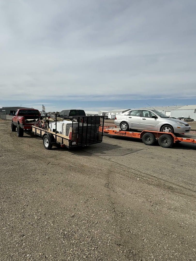 Red truck towing two trailers: one with equipment, one with a silver car, parked on gravel.
