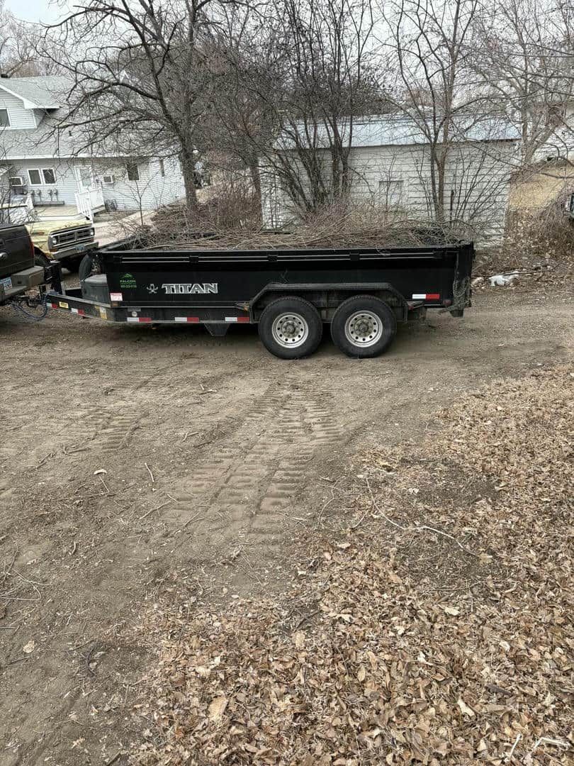 Black dump trailer parked on a dirt lot, in front of a house and trees.