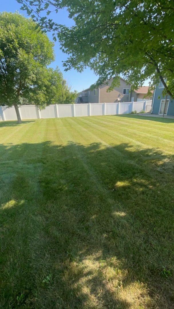 A green lawn surrounded by a white fence and trees, shadows cast across the grass.