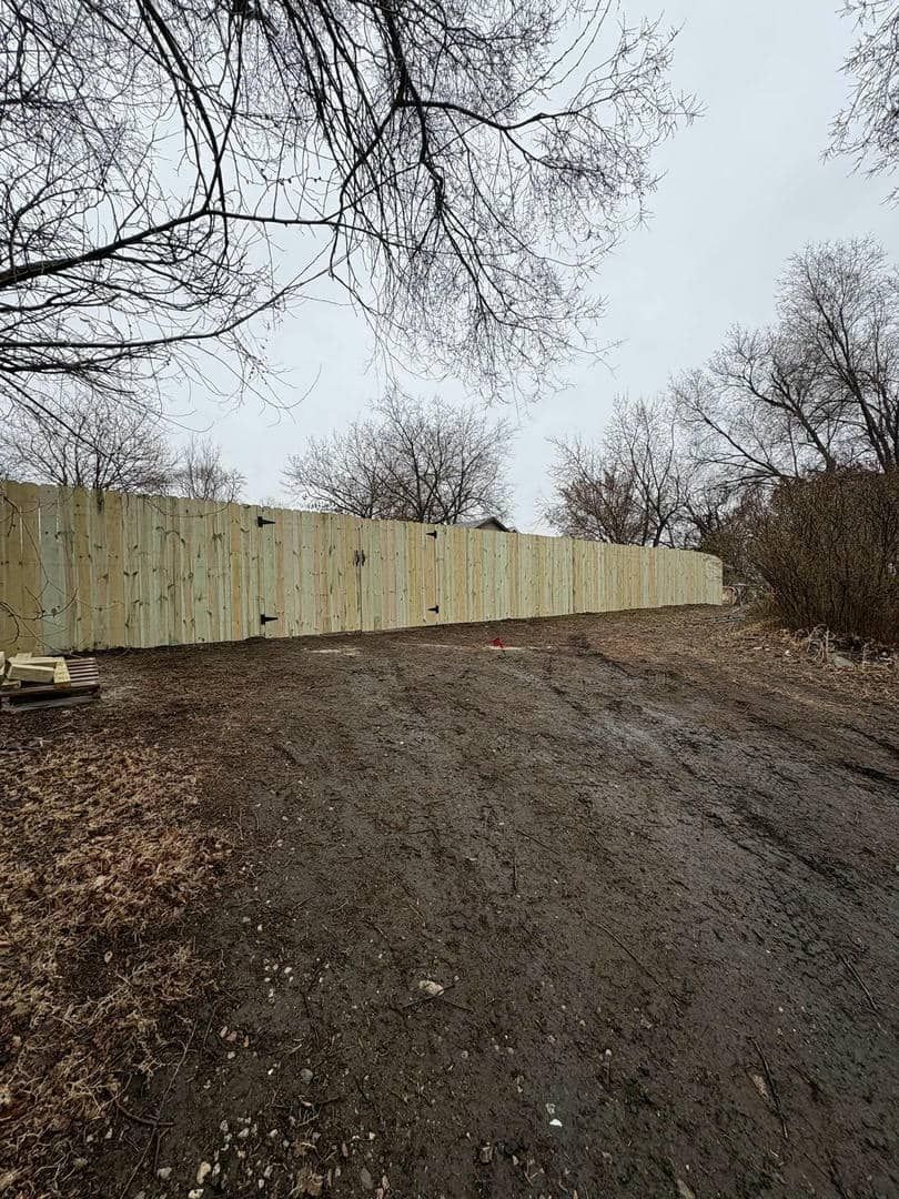 Wooden fence along a muddy hillside, with bare trees overhead on a cloudy day.