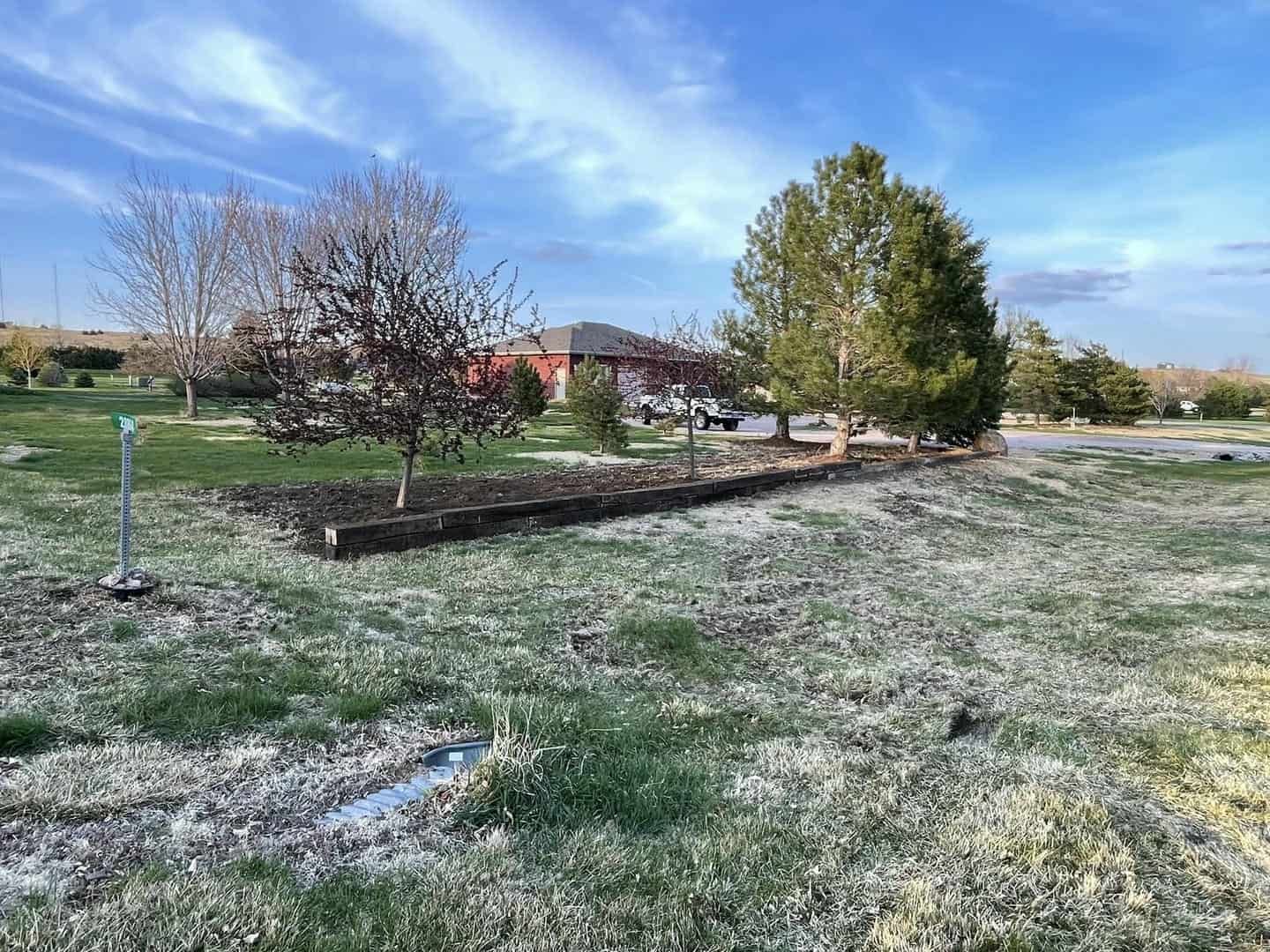 A grassy yard with bare trees, a raised garden bed, and a house under a partly cloudy blue sky.
