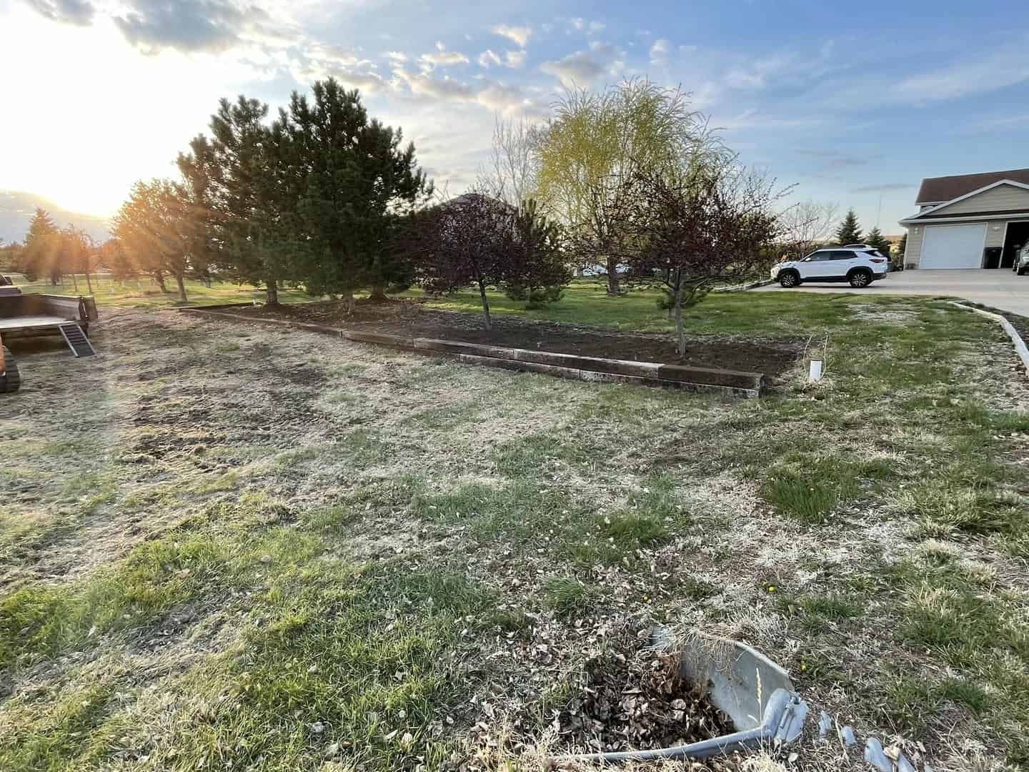 Grassy yard with raised garden bed, trees, a car, and a house under a partly cloudy sky.