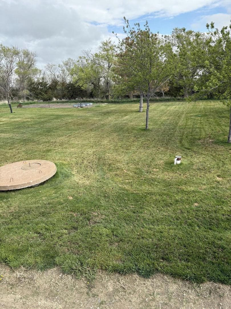 Grassy field with small trees, a circular concrete cover, and a pond visible in the distance under a cloudy sky.