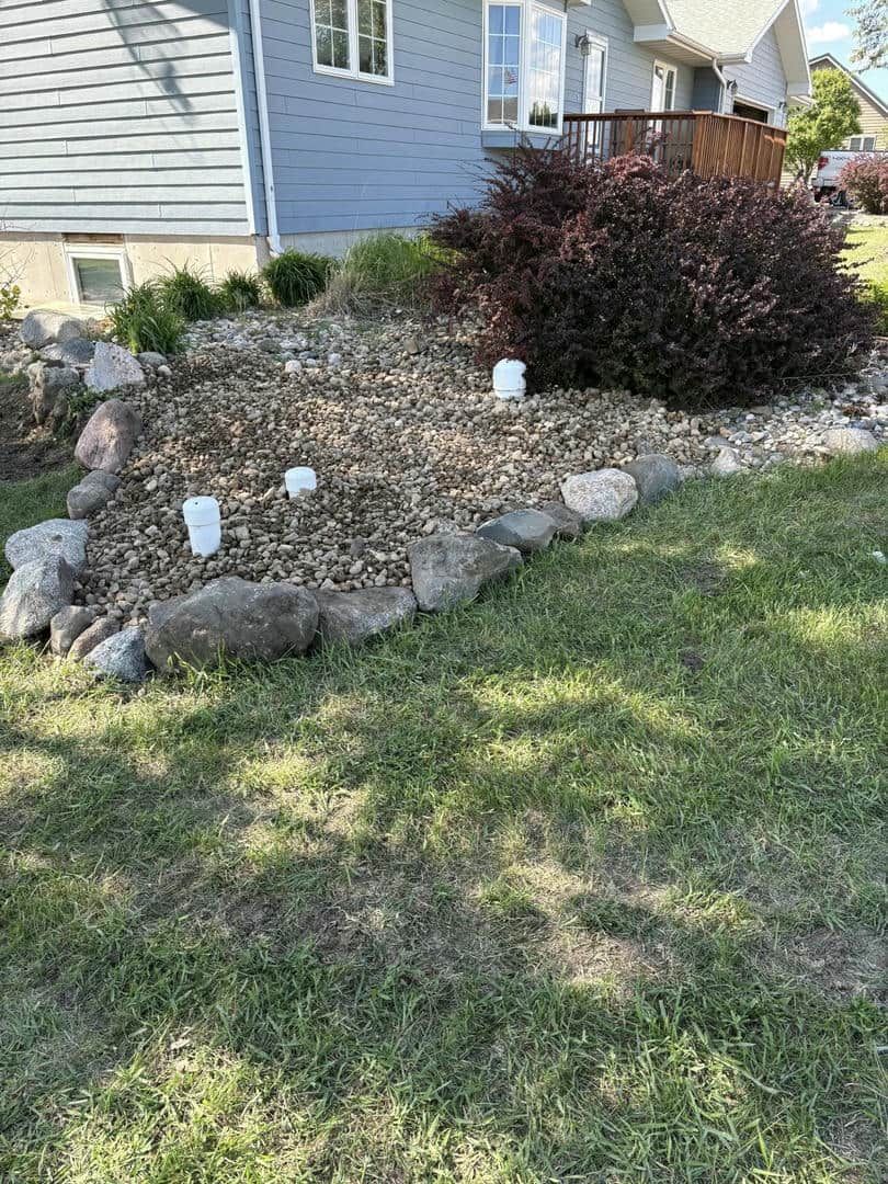 A front yard with a rock border, mulch, and bushes in front of a blue house.