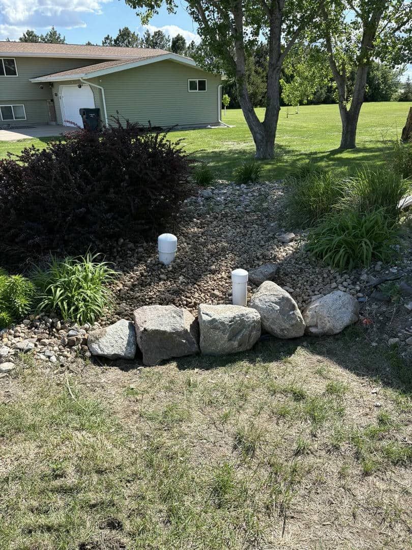White pipes in a rock-lined garden bed in front of a light green house, and lush green grass.
