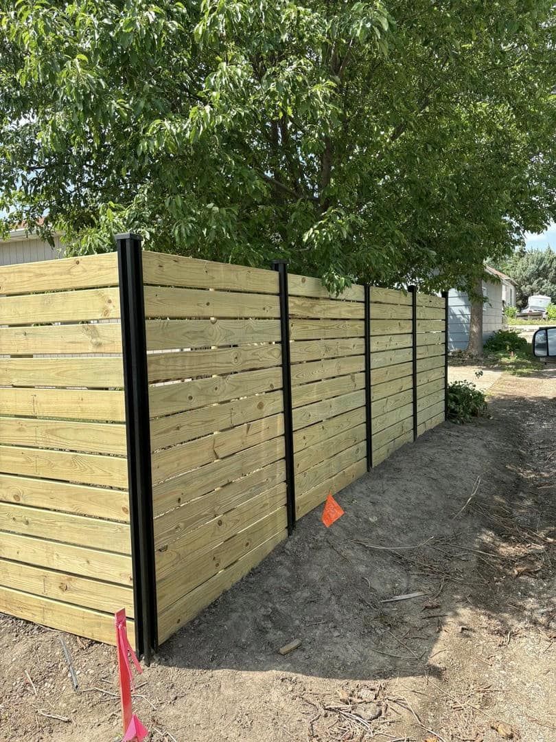 Wooden horizontal slat fence with black posts, near gravel and a tree.