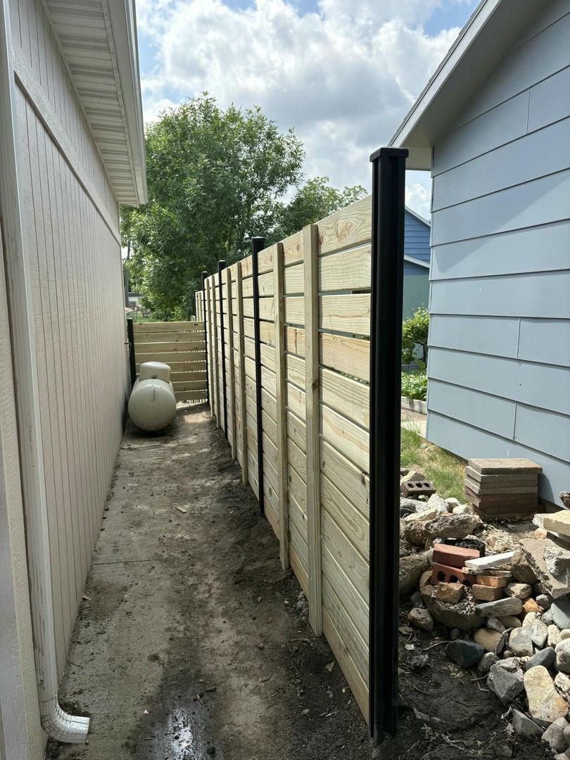 A narrow alley with a wooden fence between two buildings. The alley is dirt with construction debris.