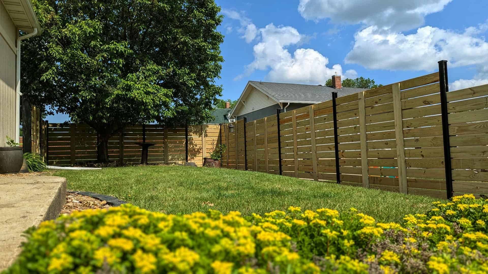 Backyard with wooden fence, green lawn, tree, and yellow flowers under a blue, cloudy sky.