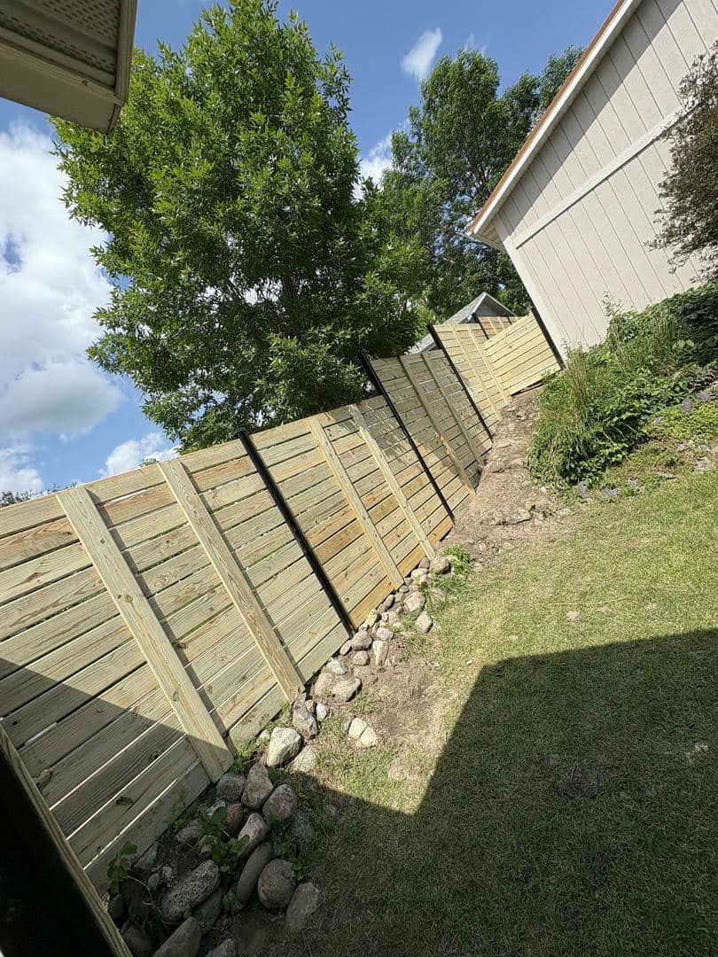 Wooden fence in a backyard with grass, rocks, and a tree, against a partly cloudy sky.