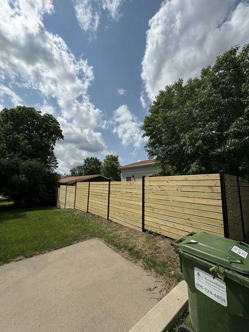 Wooden fence with woven design, green bin in foreground, blue sky with clouds.