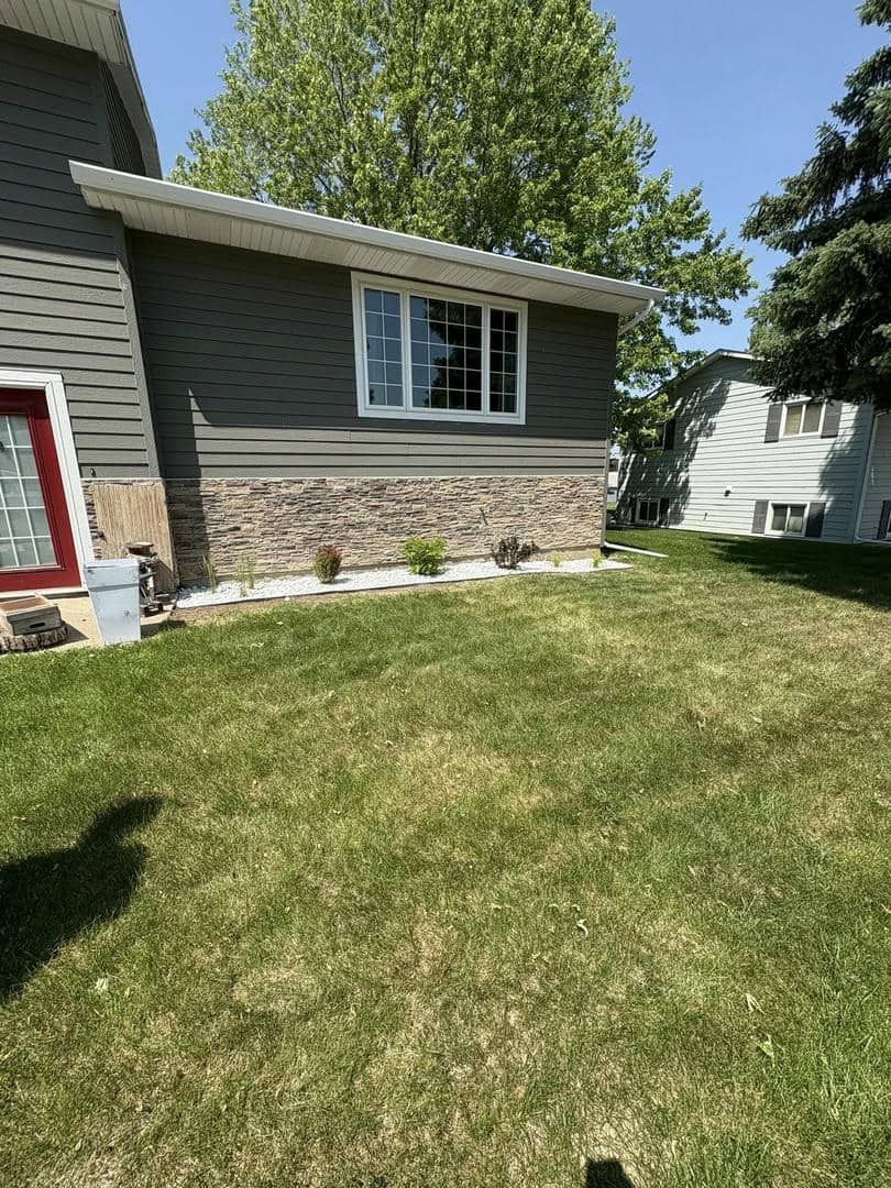 A house with green siding, a stone base, and a white-framed window, next to a grassy yard.