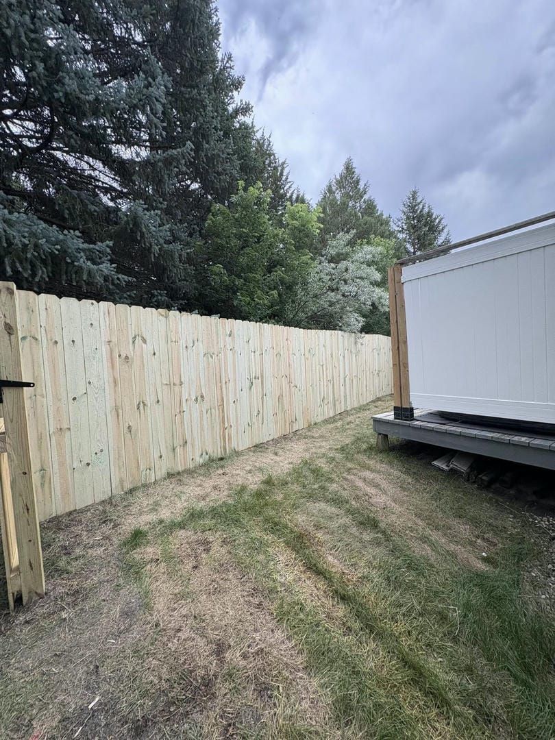 Wooden fence along a grassy yard, next to a white building; trees in the background, overcast sky.