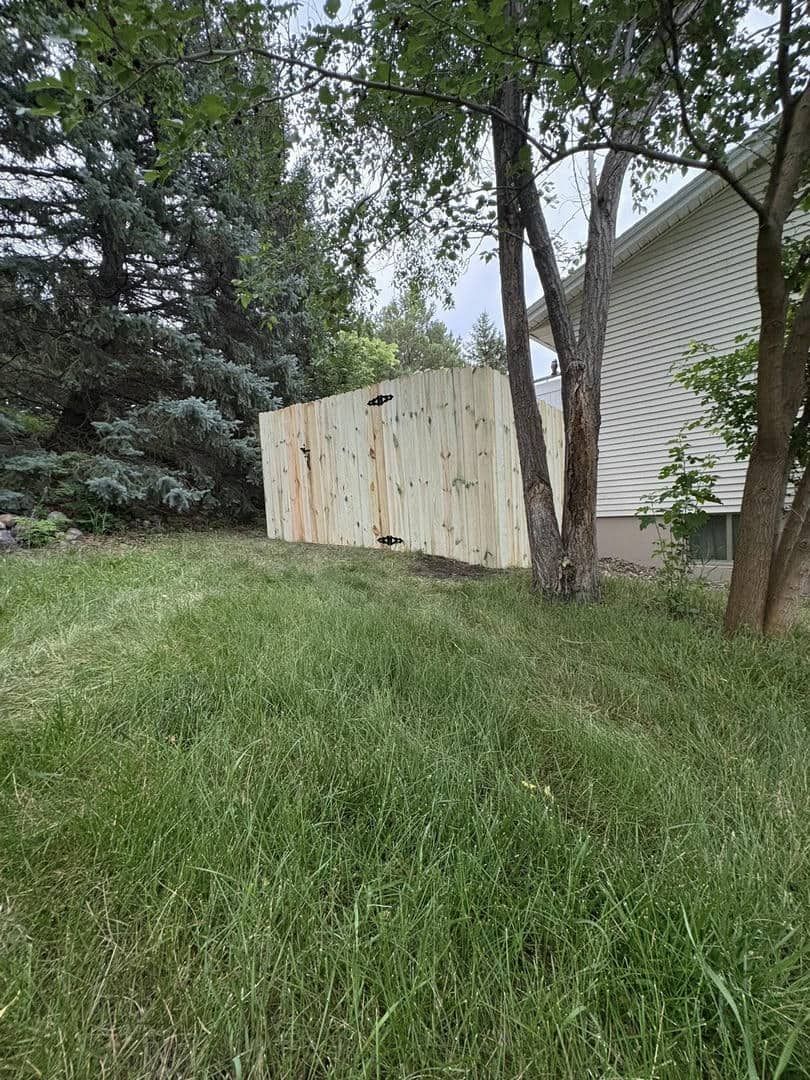 Wooden fence along a house, with tall grass in the foreground and trees in the background.