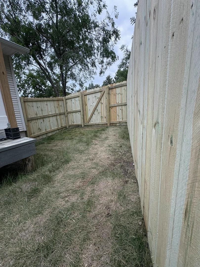 Wooden fence with gate, enclosing a grassy area, leading to trees under a cloudy sky.