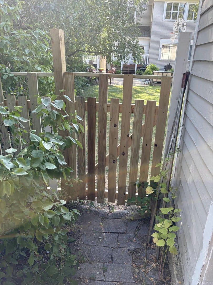 Wooden gate in a backyard, partially covered by foliage, leading to a brick pathway.