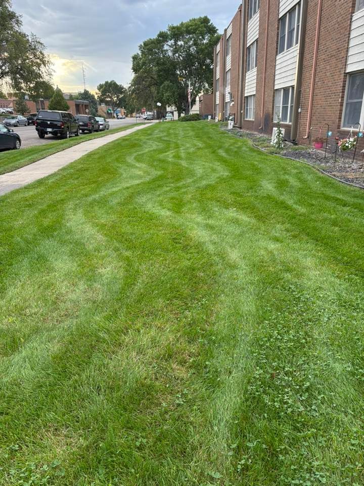 Green lawn in front of a brick apartment building with cars parked on the street.