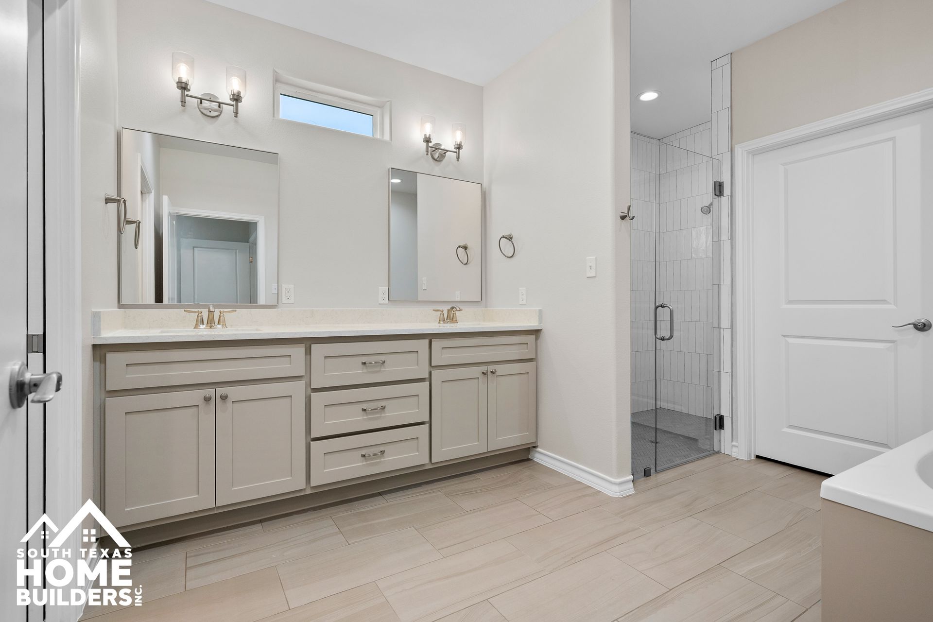 Spacious bathroom with dual sinks, shower, and closed white door. Light gray cabinets, white walls, and beige floor tiles.