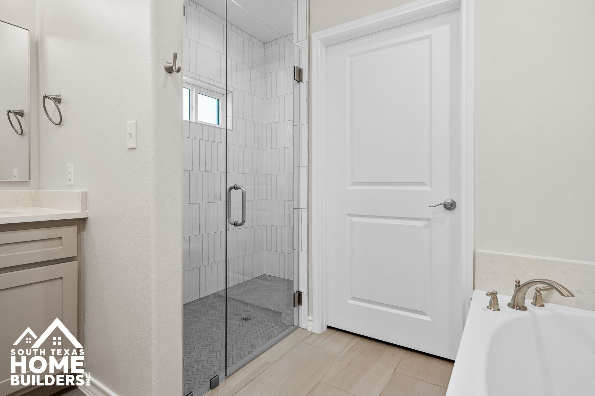 Bathroom with glass shower, white door, vanity, and bathtub. Light gray walls and light wood floors.