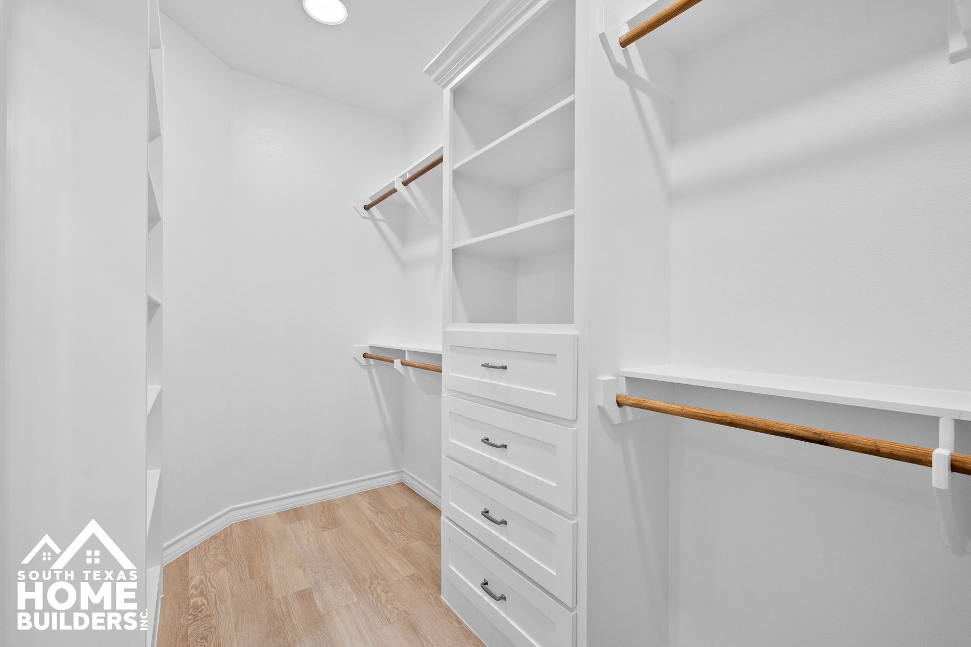 White walk-in closet with built-in shelves, drawers, and hanging rods; light wood-look flooring.
