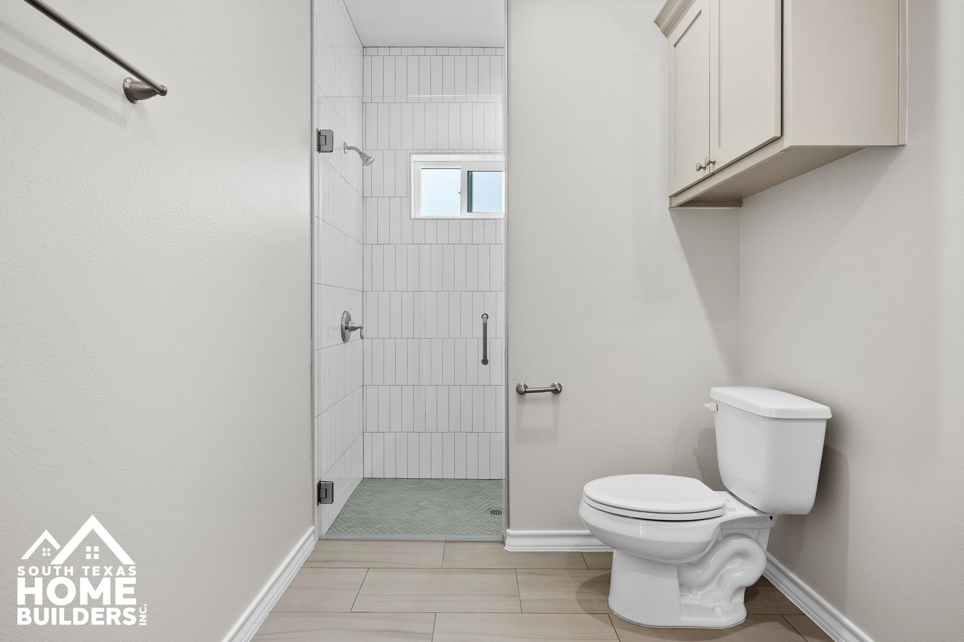 Bathroom with glass shower, toilet, and light-colored cabinets against neutral walls and tiled floor.