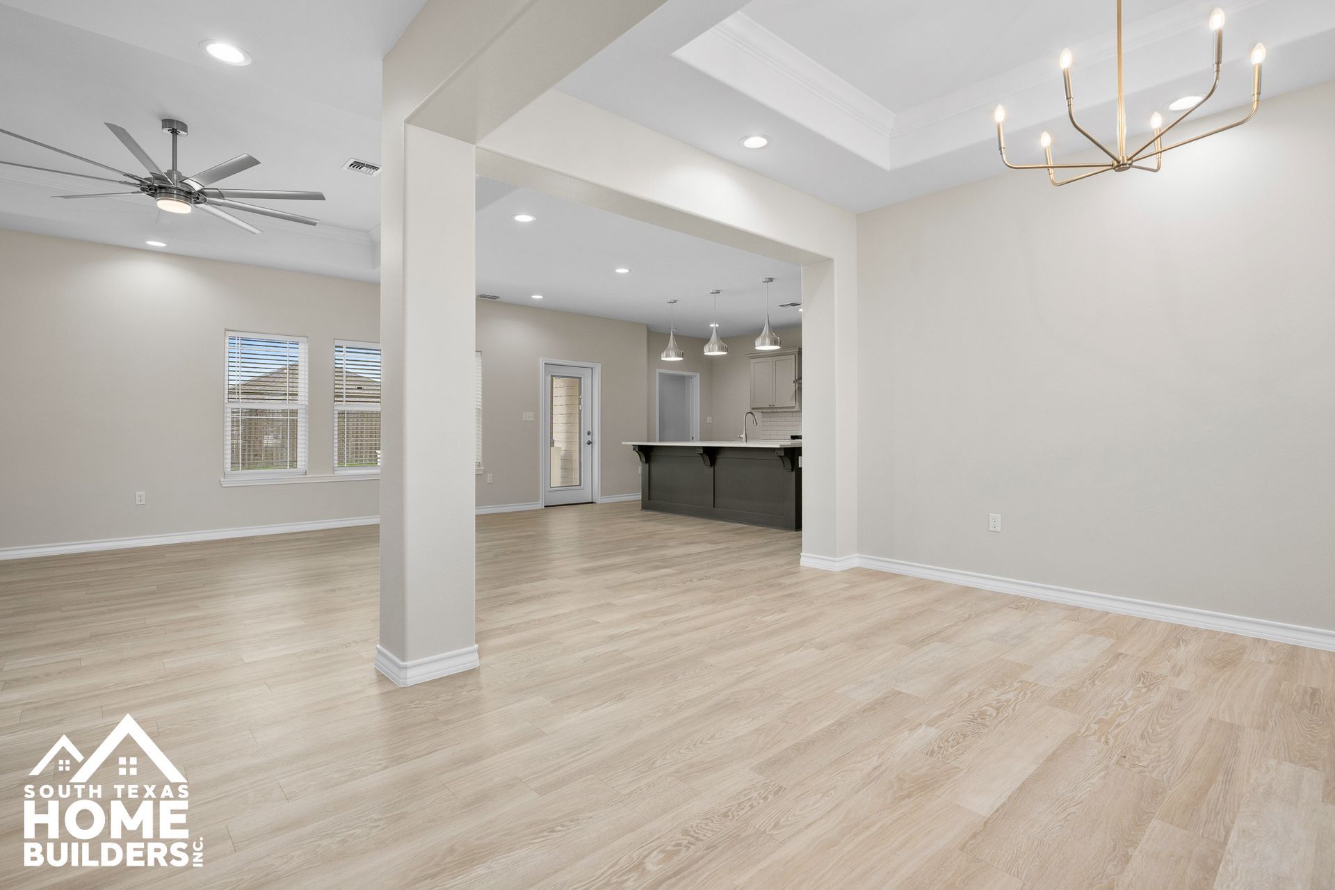 Empty interior room with light wood floors, columns, and a view to the kitchen.