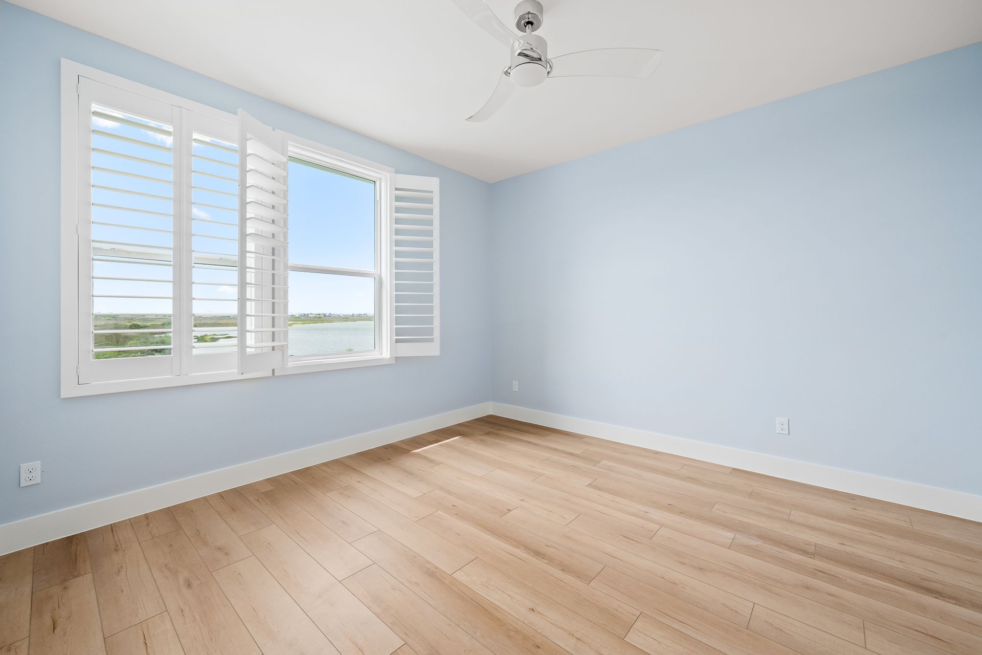 An empty bedroom with hardwood floors , white shutters and a ceiling fan.
