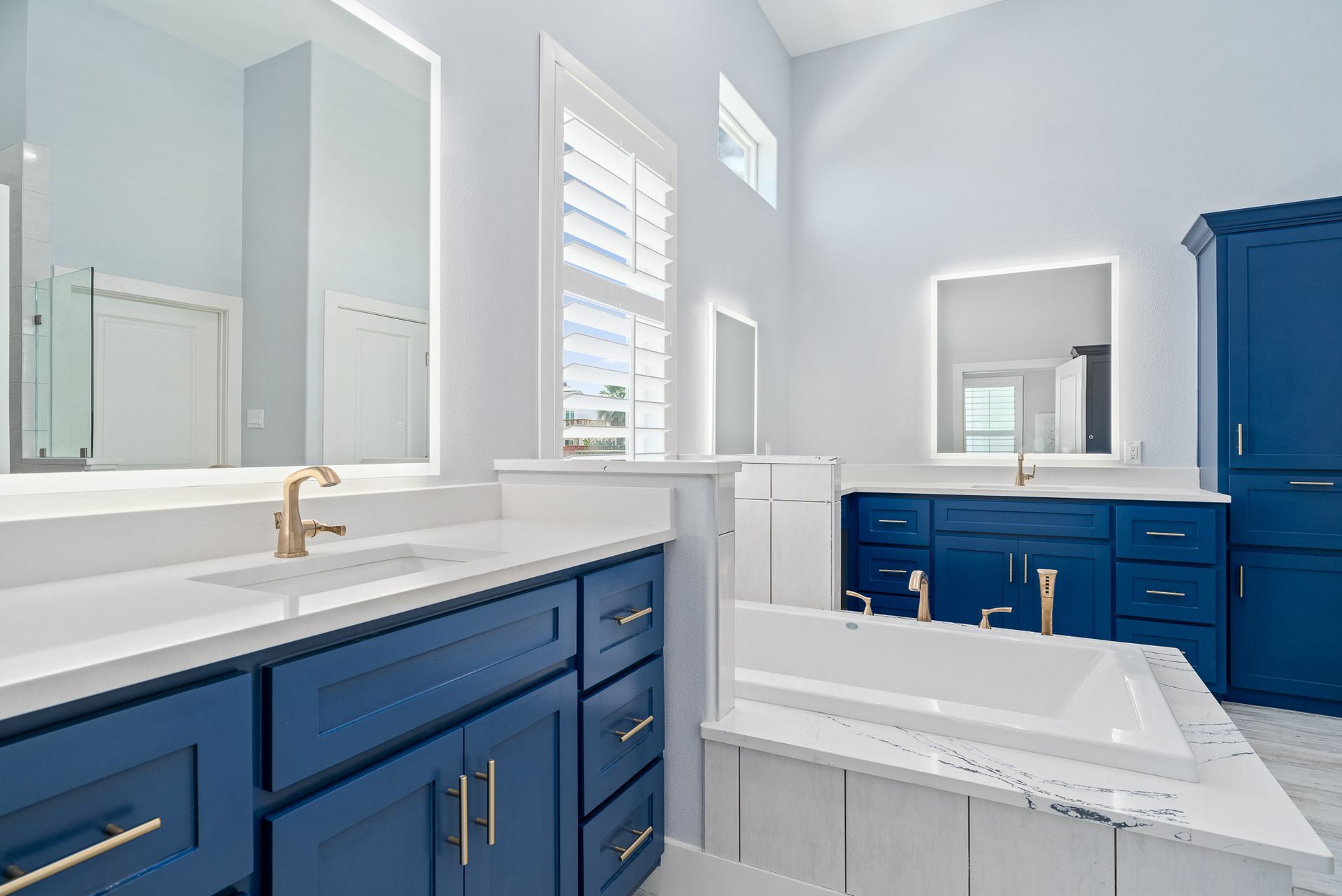 A bathroom with blue cabinets , white counter tops , and a bathtub.