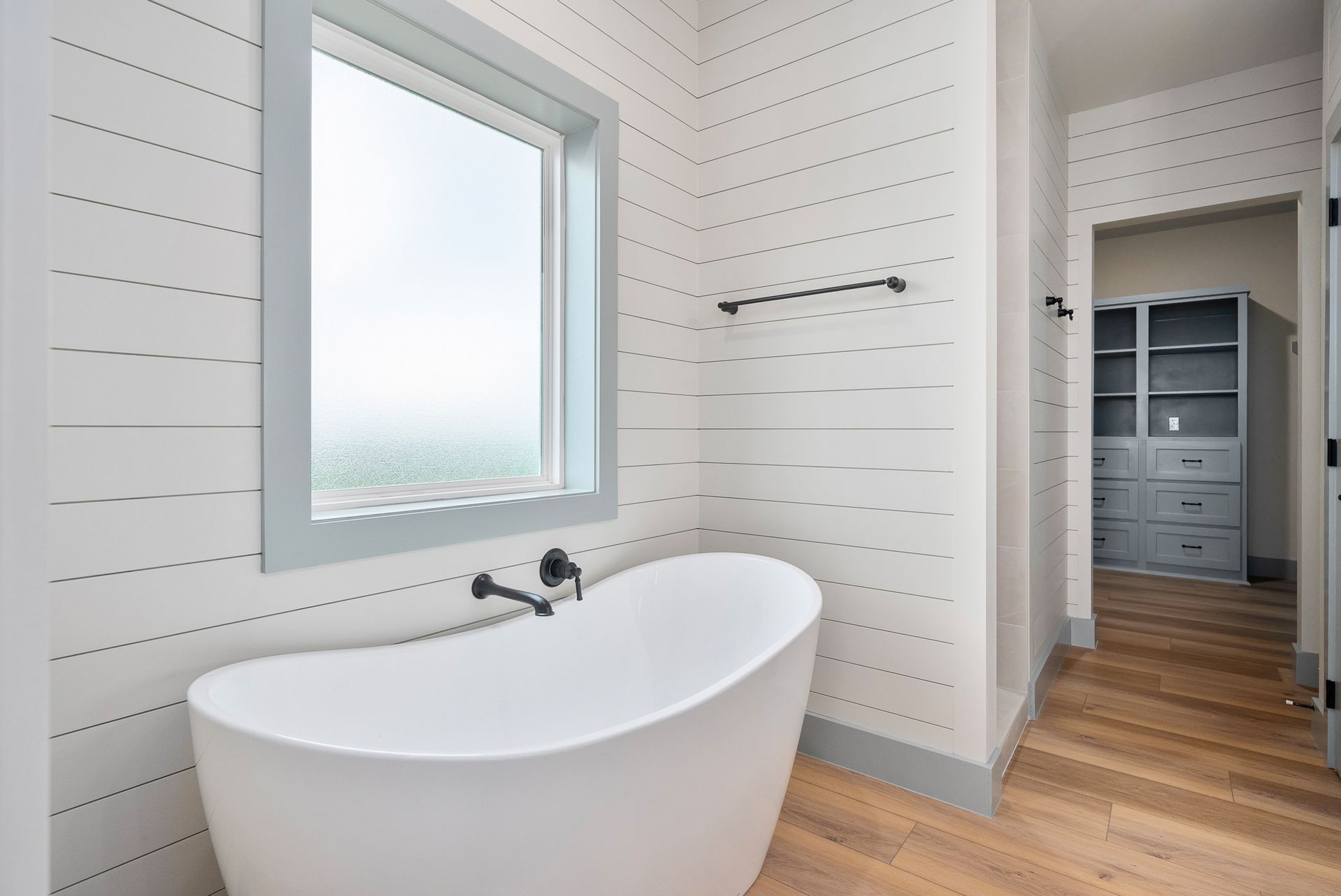White freestanding bathtub near a window in a bathroom with horizontal wood paneling and light wood flooring.