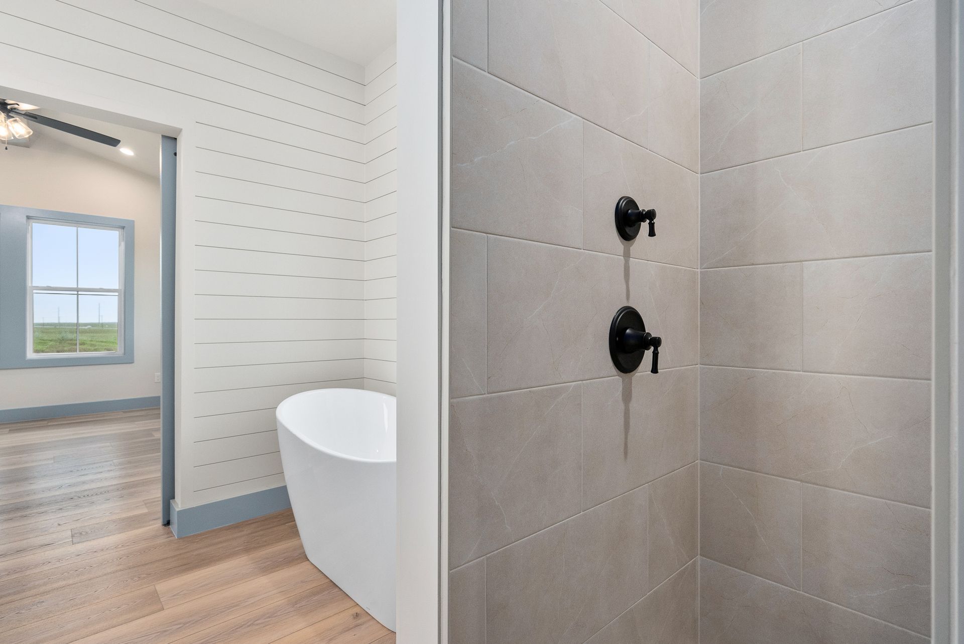 Bathroom interior with a shower, bathtub, and view of another room with a window.
